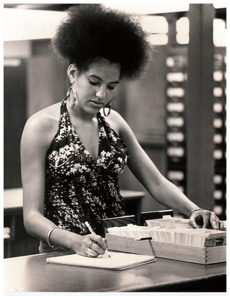 Black and white photo of a woman writing while standing in the library